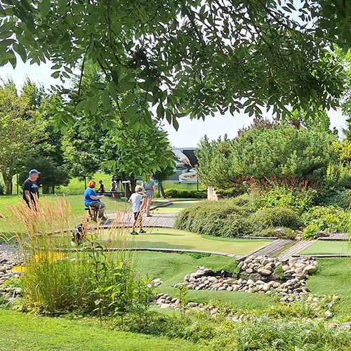 Rasen und Steinflächen mit Personen die auf einer Bank sitzen oder auch welche die Abenteuergolf spielen mit Büschen und Bäumen. Im Hintergrund sind Gebäude vom Erlebnsipark und dem Ocean wave zu sehen.